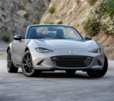 A silver MX-5 ST drives down a road cutting through the mountains.