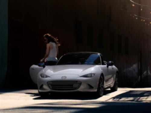 A person rests one hand on an MX-5 ST near the ocean while looking out at the horizon.