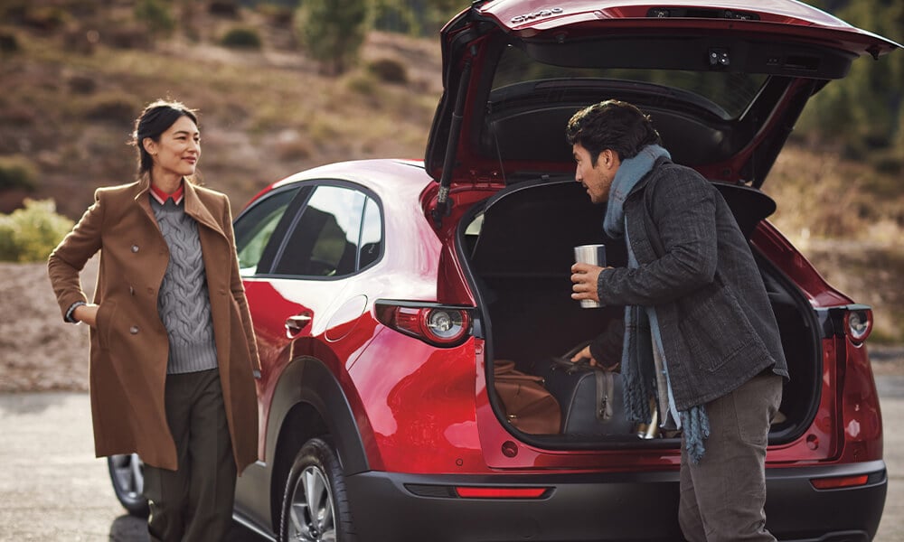 n homme et une femme se tiennent près du coffre d’un Mazda CX-30 rouge vibrant cristal métallisé avec un paysage extérieur flou en arrière-plan.