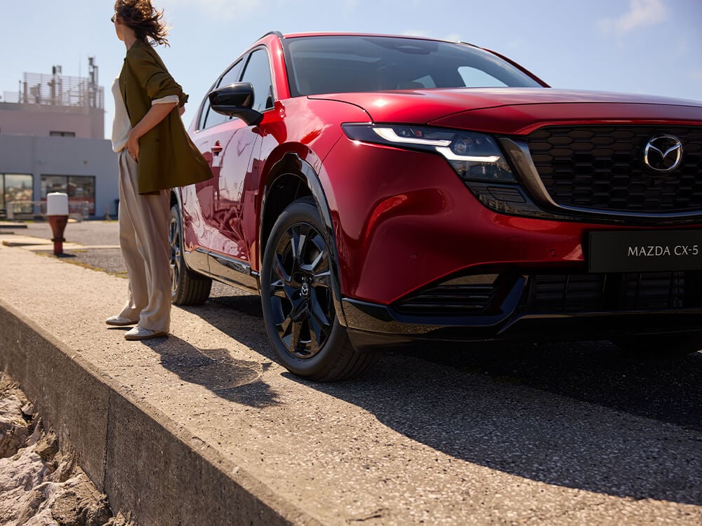 Front corner low angle of a Soul Red Crystal Mazda CX-5 parked, with a women standing beside the passenger side, looking away from the camera.