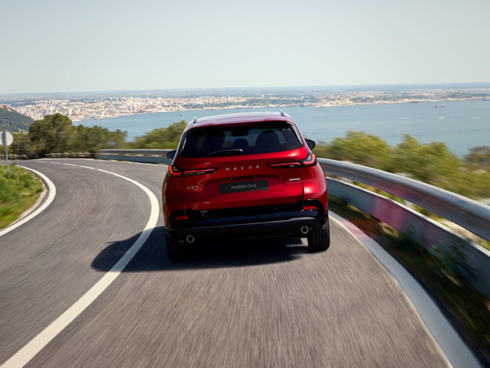 Rear view of a Soul Red Crystal Mazda CX-5 turning on an elevated road, with a blurred town visible in the distance near a body of water.