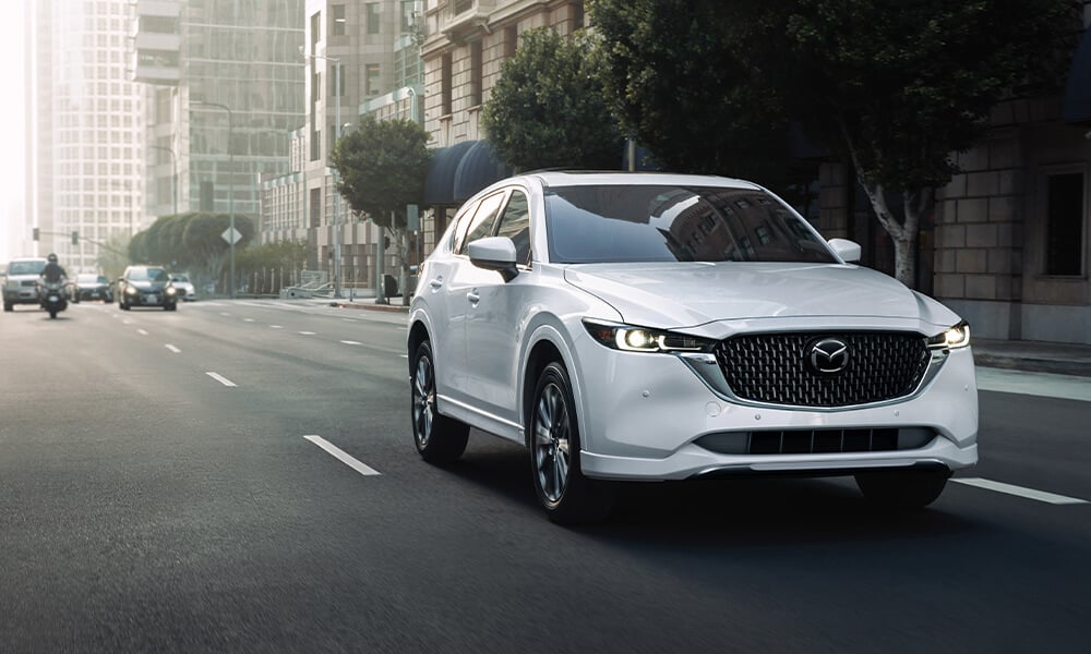 Front side profile of a Rhodium White Metallic Mazda CX-5 driving towards the camera on a city road.