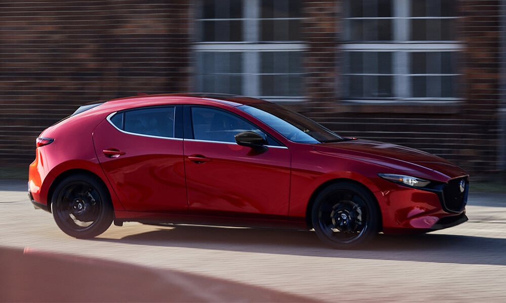 Side profile of a Soul Red Crystal Metallic Mazda3 Sport driving past a blurred building.
