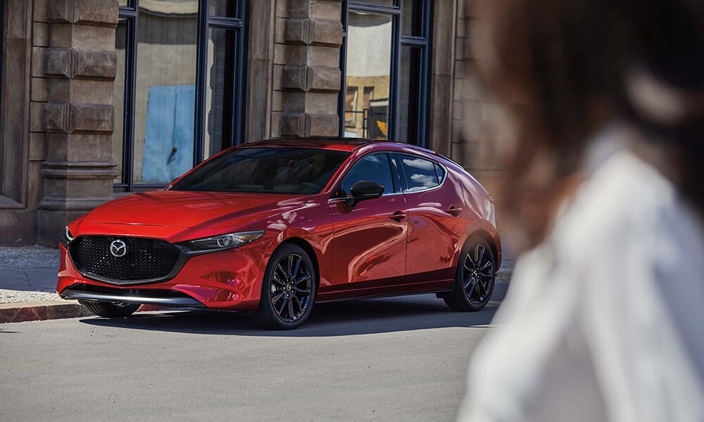Blurred woman in the foreground looking towards a parked Soul Red Crystal Metallic Mazda3 Sport shown in side profile.