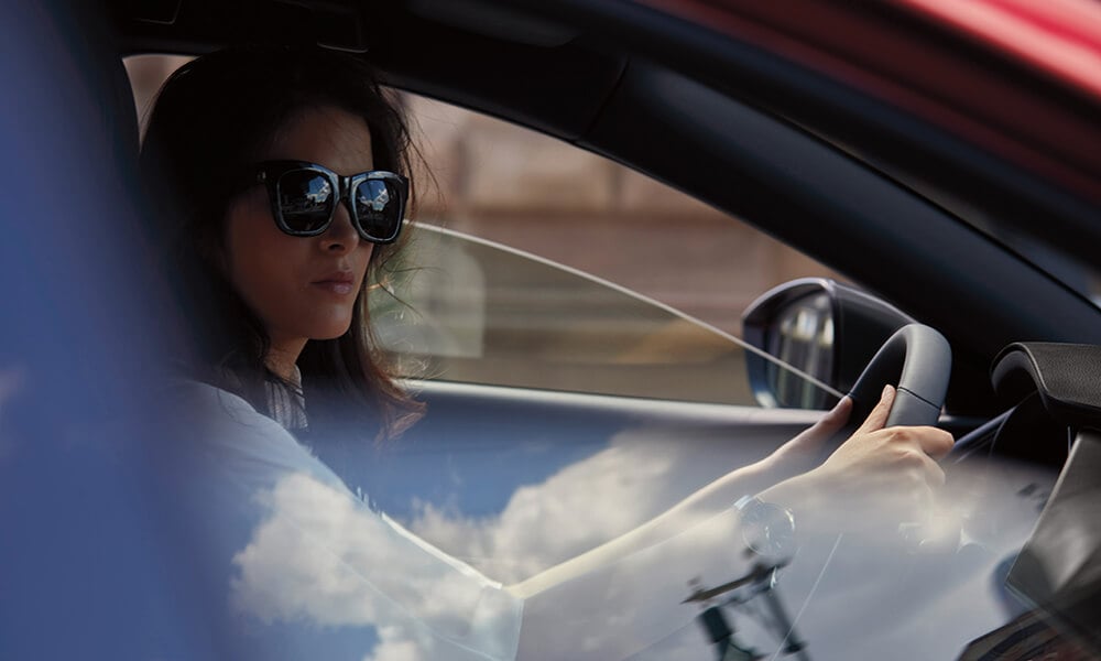 Side profile of a woman wearing sunglasses, seen through the open passenger-side window, driving a Mazda3 Sport.