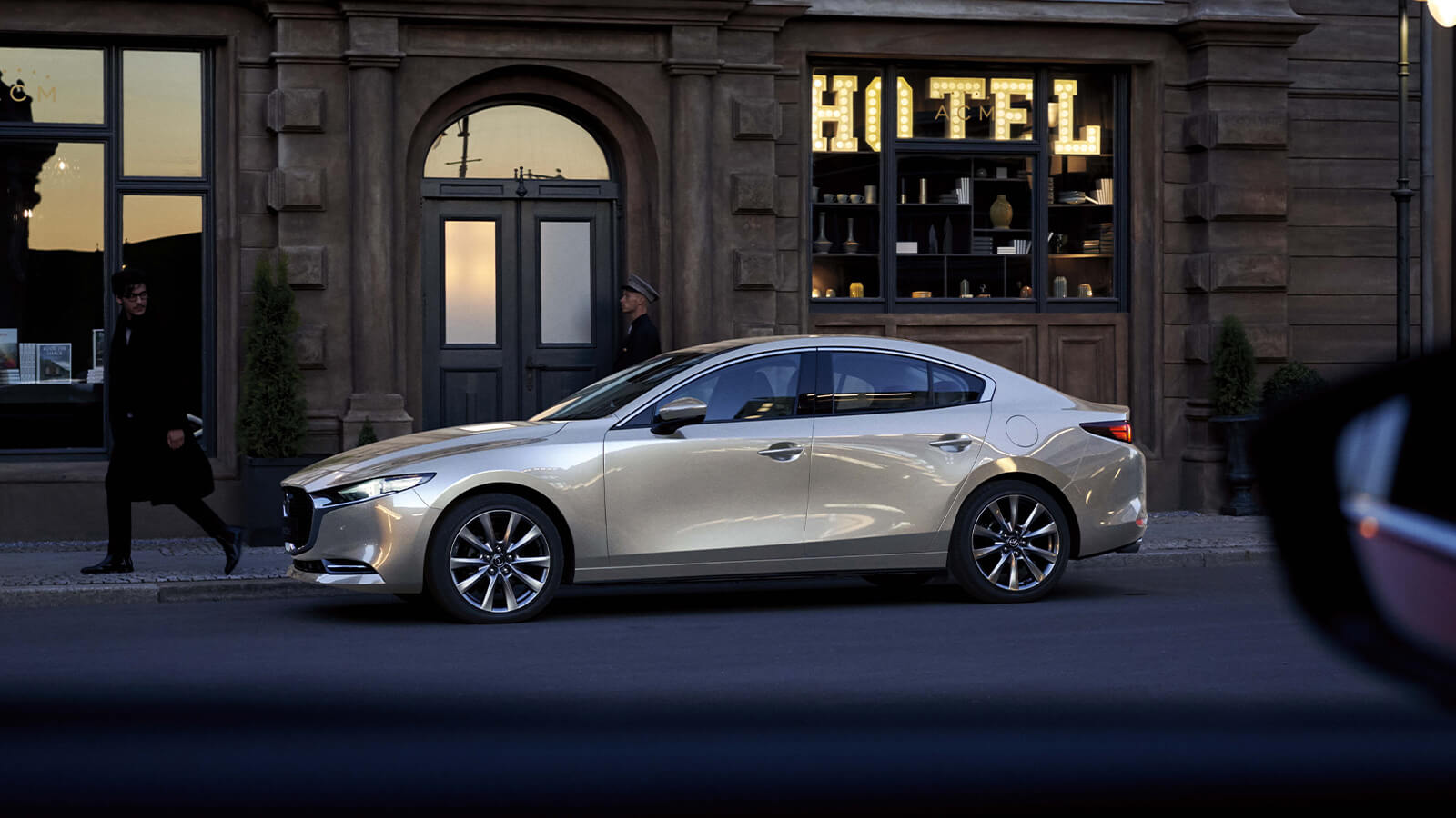Vue de profil d’une Mazda3 quartz platine métallisé stationnée à l’avant d’un immeuble.
