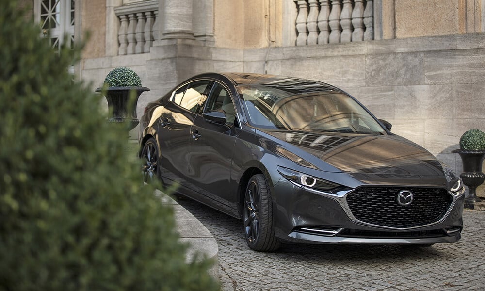 Vue de l’avant d’une Mazda3 gris mécanique métallisé avec un buisson en pot décoratif à l’avant et un manoir à l’arrière.